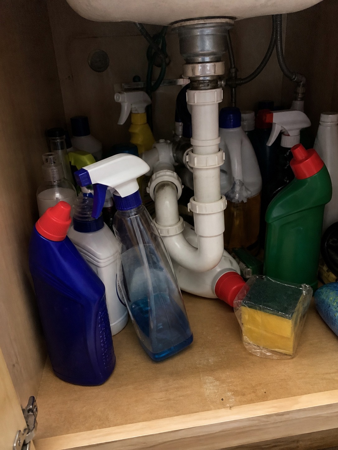 Cluttered under-sink cabinet with cleaning bottles fallen around plumbing pipes