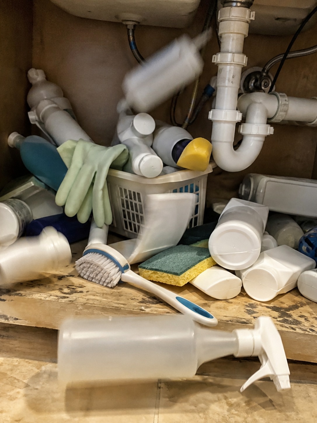 Cluttered under-sink cabinet with damp cleaning bottles, crowded pipes, and a dark back corner